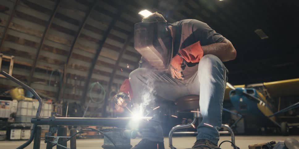 Man welds a project while in a hanger holding a yellow and blue airplane