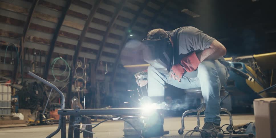 A person wearing a welding helmet is welding a metal object and making several white sparks come out