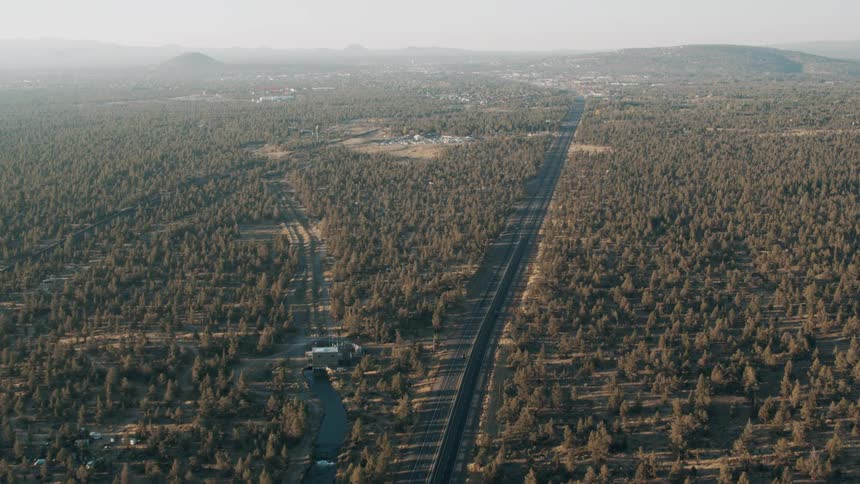 A long straight highway cuts through a vast forested landscape with distant hills and a small town visible on the horizon