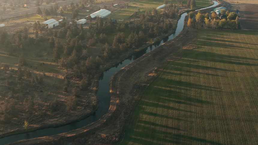 An aerial view of a small river running through grassy fields with some trees and some houses on the banks