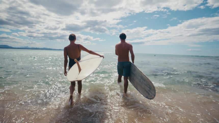A man and woman are carrying surfboards while walking through ocean water on a partly sunny day