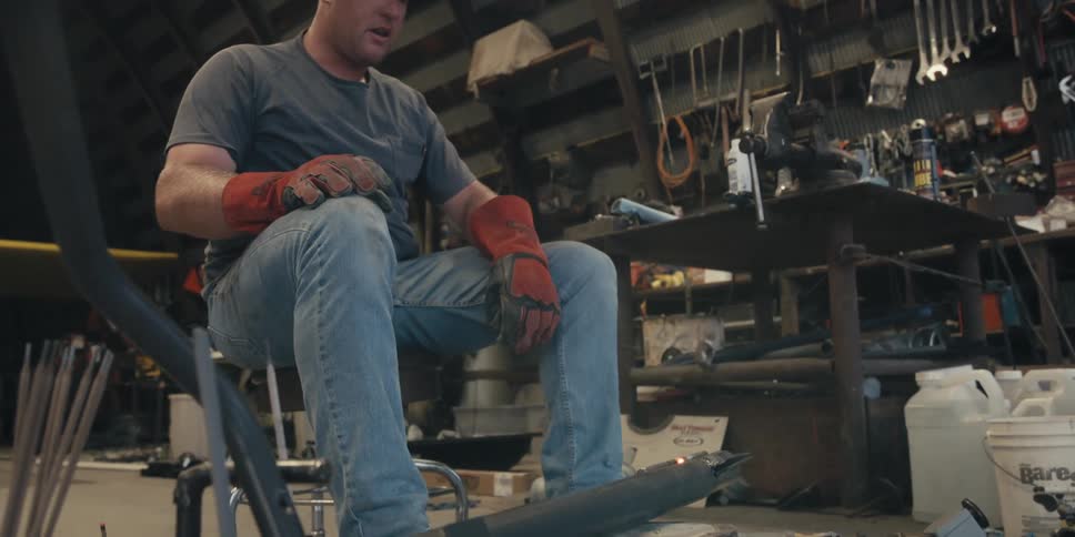 A man sitting in a chair wearing gloves and a helmet to weld a piece of iron and then a man crouches in front and looks