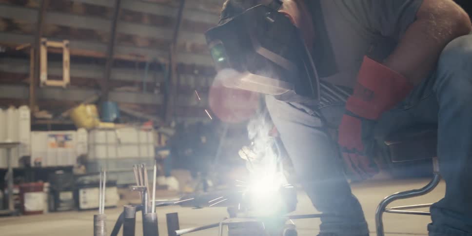 Worker man wearing a helmet and protective gear sitting down on a stool indoors soldering a piece of metal