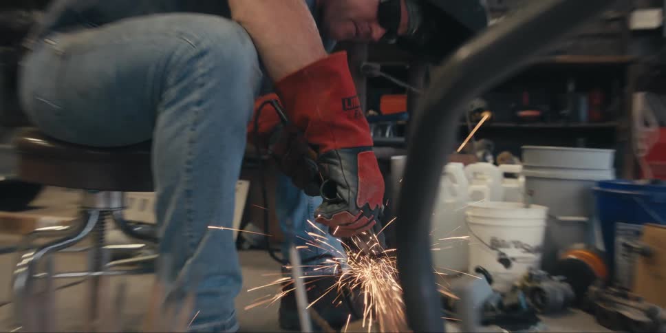 A man sitting on a chair wearing a helmet and sunglasses welding a metal object and is giving off several sparks