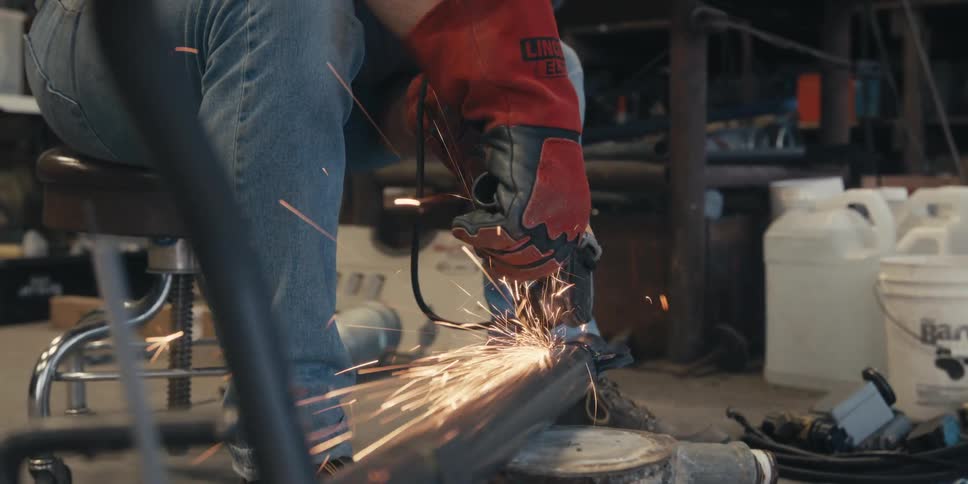 A person wearing red protective gloves is using a power tool to grind a metal pipe producing bright sparks in a workshop setting