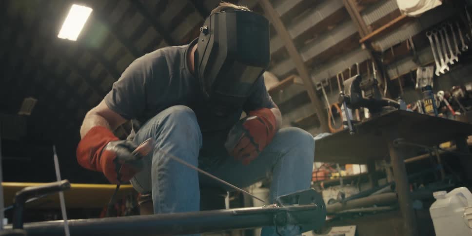 A crouched man wearing a welding helmet is welding a piece of iron that is in front of him and giving off several sparks