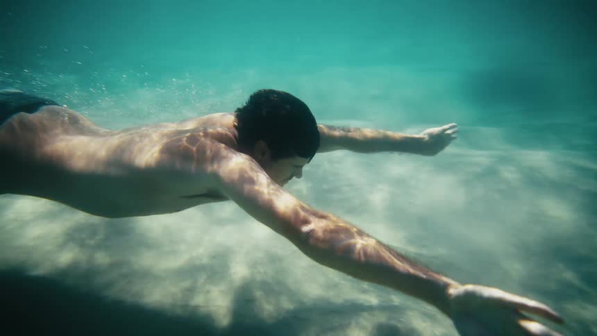 A young shirtless man diving underwater in clear water with some lights coming from the surface
