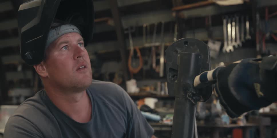 A man with a welding helmet on his head is looking at a metal tool while a person with a glove is pointing at the tool