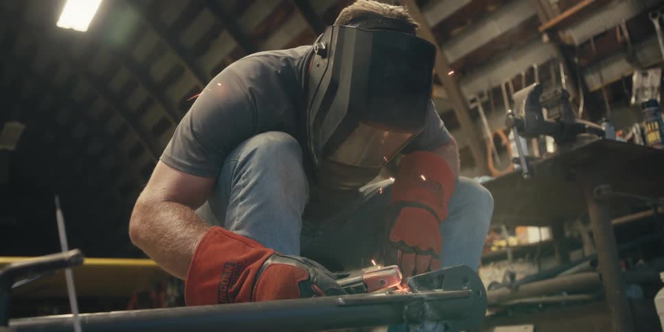 A man crouching down wearing a helmet to weld a piece of iron and making several sparks jump upwards