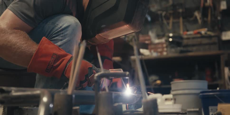 A person wearing protective gear including a welding helmet and orange gloves is welding metal in a workshop
