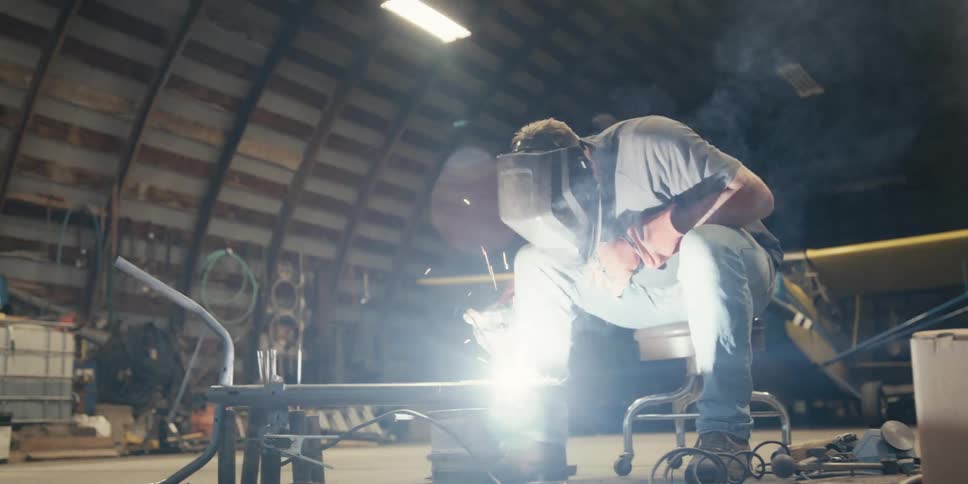 The man wearing the welding helmet is using gloves to weld a steel beam in front of him and is giving off several sparks
