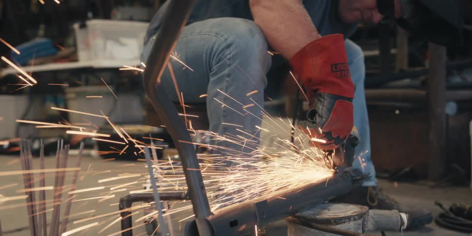 A person wearing red protective gloves is using a power tool to grind a metal pipe producing bright sparks in a workshop setting