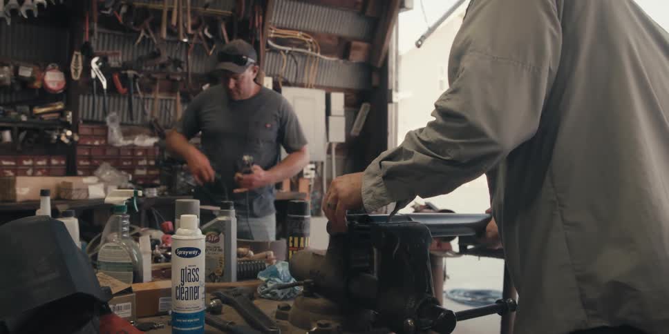 A person with his back turned is clamping a metal object in a vise that is clamped to a table in a workshop