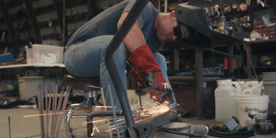 A person wearing red protective gloves is using a power tool to grind a metal pipe producing bright sparks in a workshop setting