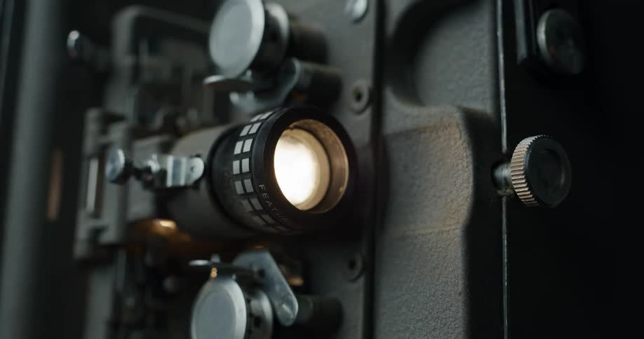 A closeup view of an oldfashioned film projector with a lit lens and various adjustment knobs and levers