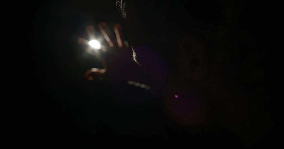 A hand reaches out towards a bright light source in a dark room casting a shadow on a face
