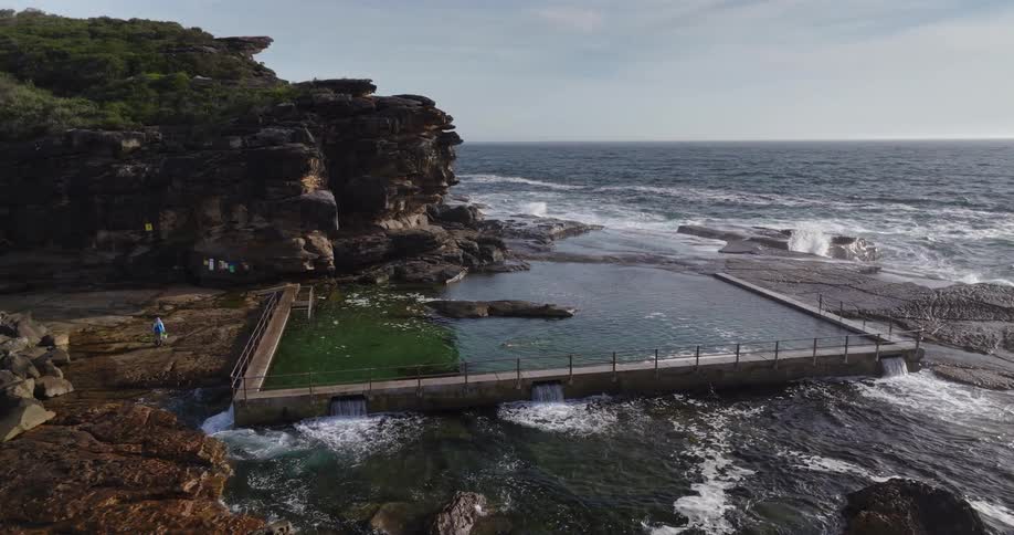 Swimmer swims in a coastal pool near a forest covered cliff while a person carries fishing equipment nearby