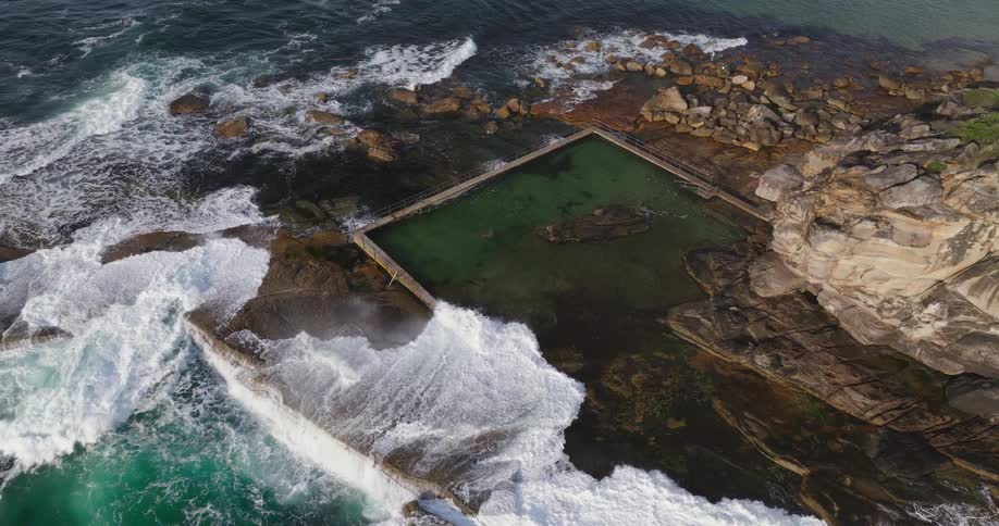 An aerial view shows ocean waves crashing against the rocky edges of a rectangular sea pool with swimmers inside