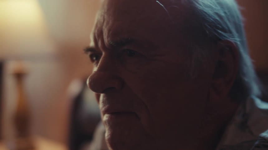 An elderly person with white hair is sitting indoors wearing a striped shirt with soft lighting illuminating the scene