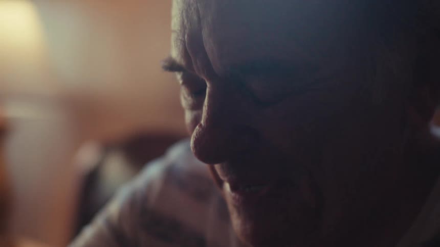 An elderly person with white hair is sitting indoors wearing a striped shirt with soft lighting illuminating the scene
