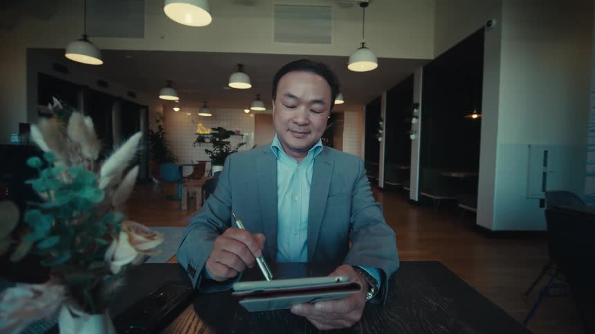 A seated man wearing formal wear is typing on a tablet using a pen and has a small smile on his face
