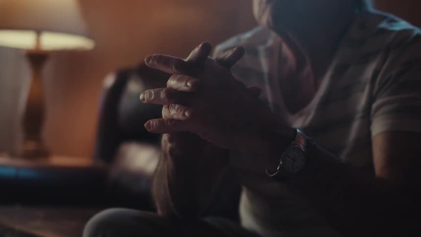 An elderly person with white hair is sitting indoors wearing a striped shirt with soft lighting illuminating the scene