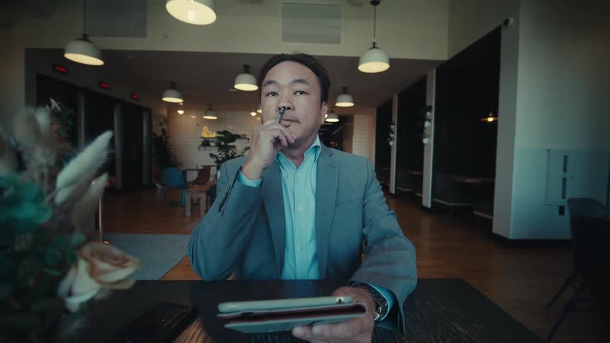 A man is deep in thought and occasionally uses his stylus on his tablet as he sits at a desk