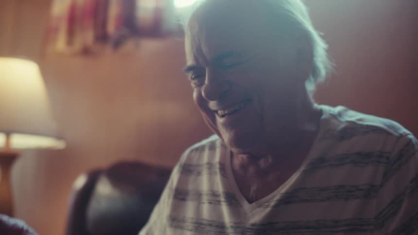 An elderly man sits on a dark leather couch at home smiling and laughing while using a tablet