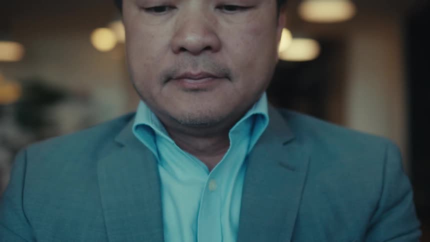 A man in formal wear is sitting and fiddling with a book in front of him and he is talking on the phone while putting his finger on his head to think