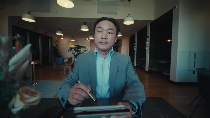 A person in a suit is sitting at a table in a modern house holding a tablet and a stylus with a vase of dried flowers