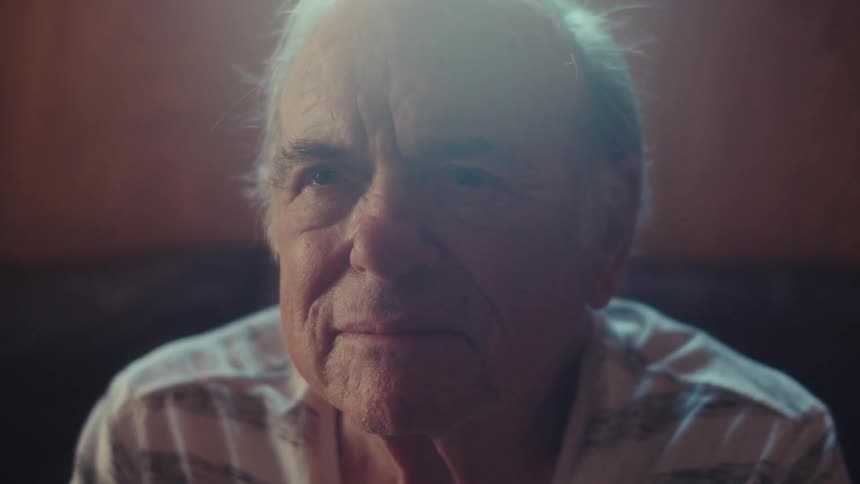 An elderly person with white hair is sitting indoors wearing a striped shirt with soft lighting illuminating the scene