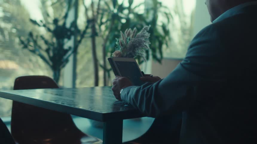 A person in a suit is sitting alone at a table in a modern plantfilled room using a tablet with a stylus