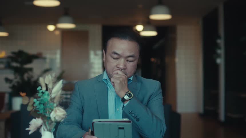 A person in a blue suit is resting their chin on their hand while working on a tablet