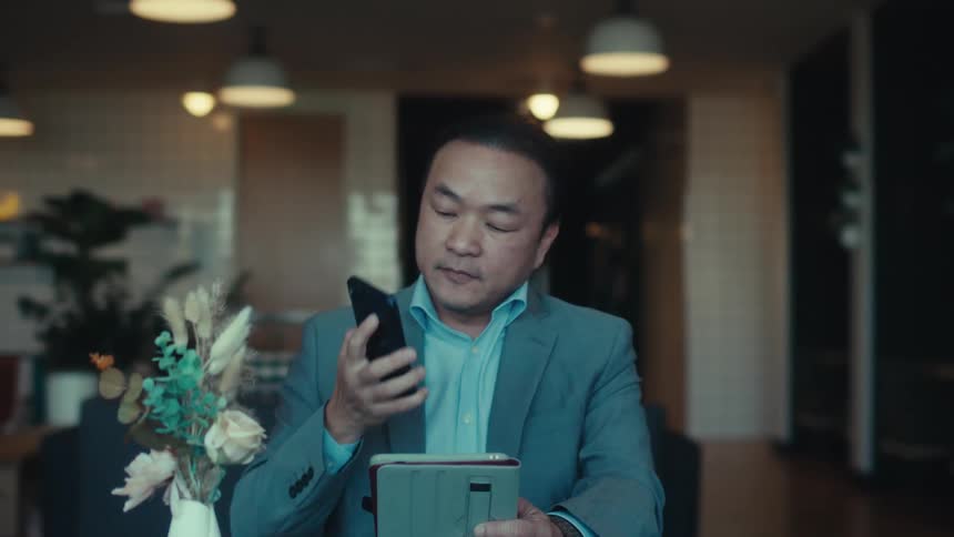 A person in a suit is sitting at a table in a modern stylish caf with a tablet answering a call on his phone