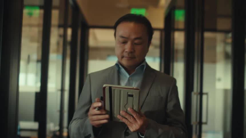 A person in a suit is walking down a modern office hallway while writing on a tablet