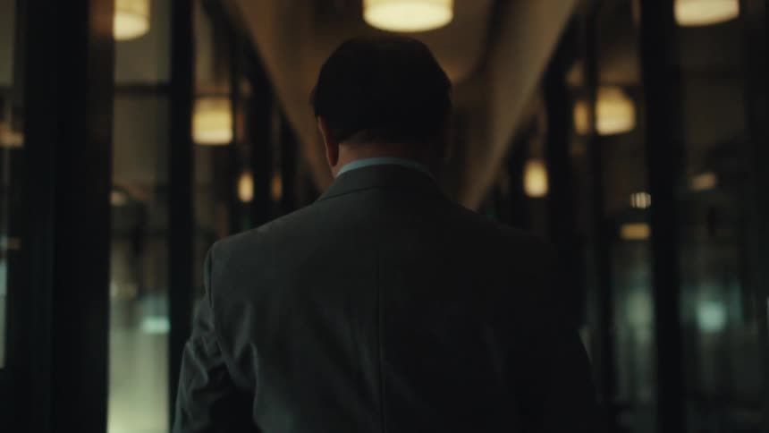 A man in a suit walks down a dimly lit corridor of a modern office seen from behind