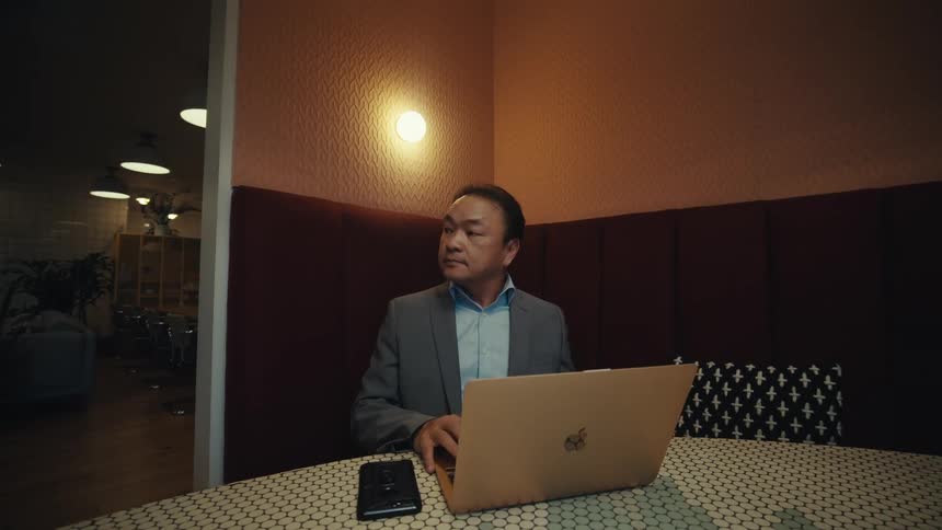A man in a suit is shown talking on his phone working on his laptop and then closing it while seated at a table