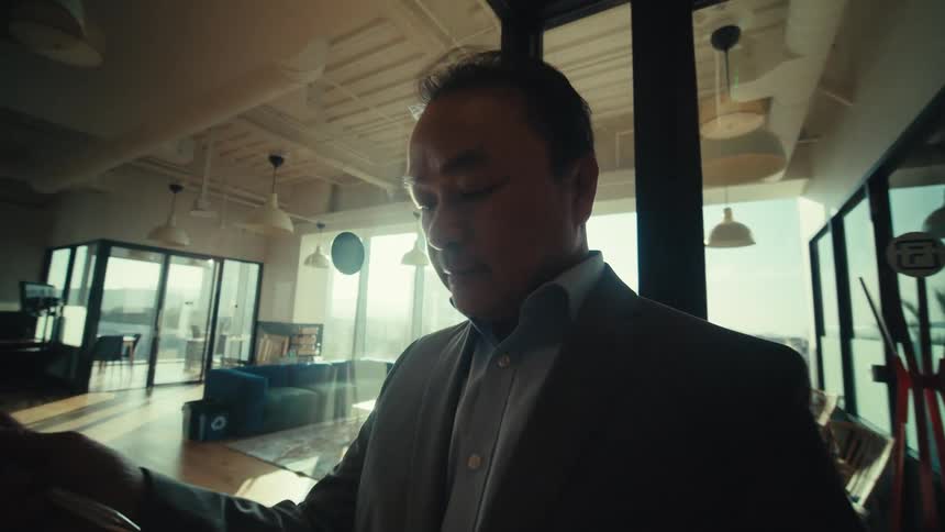 A man in formal clothes walking into a glasswalled hallway and he is fiddling with a device in his hands
