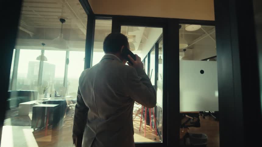 A person in a suit is walking down a modern office hallway while talking on a smartphone
