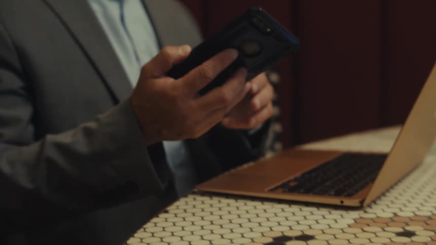 A person typing on a laptop that is on a table then picks up a cell phone that is next to it