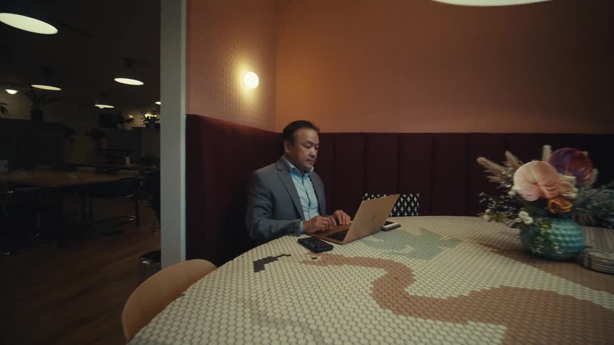 A man wearing a suit is shown working on a laptop at a round table in a modern office setting