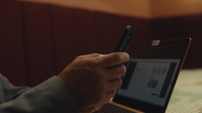 A person sitting at a table with an open laptop picks up a smartphone