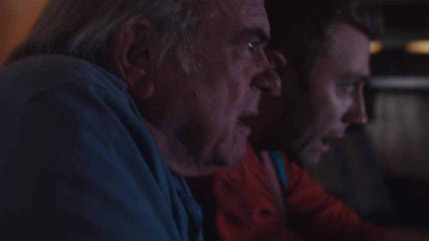 Father and son are sitting on a dark leather couch in a dimly lit living room playing video games
