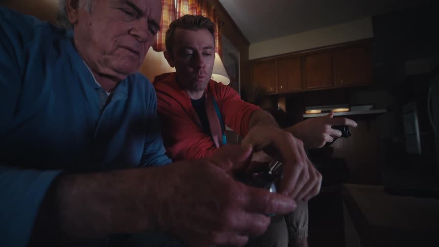 Father and son are sitting on a dark leather couch in a dimly lit living room playing video games