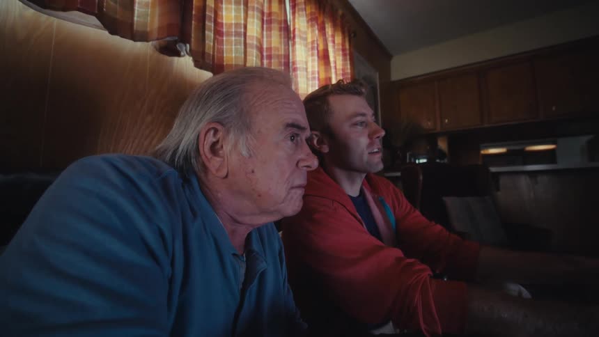 Father and son are sitting on a dark leather couch in a dimly lit living room playing video games