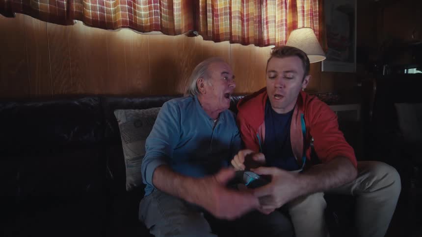 Father and son are sitting on a dark leather couch in a dimly lit living room playing video games