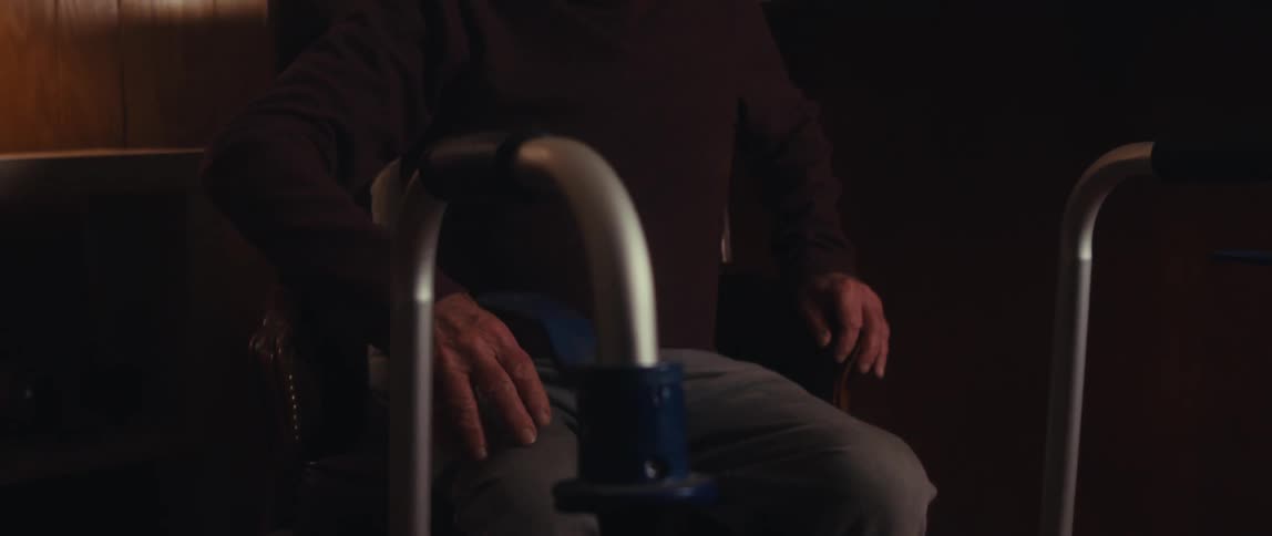 An elderly man uses a walker for support as he sits down on a chair in the living room