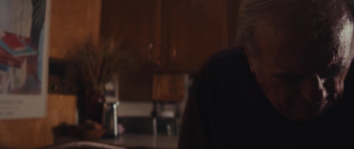 Elderly man walks slowly across the room to sit in a chair near the kitchen during the day