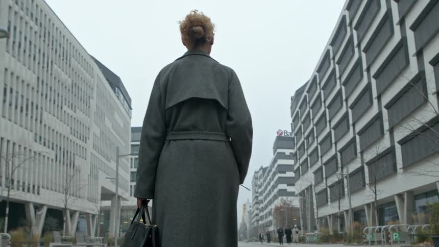 A woman with curly hair wearing a long gray coat and carrying a black bag is walking down a modern urban street flanked by tall geometric buildings