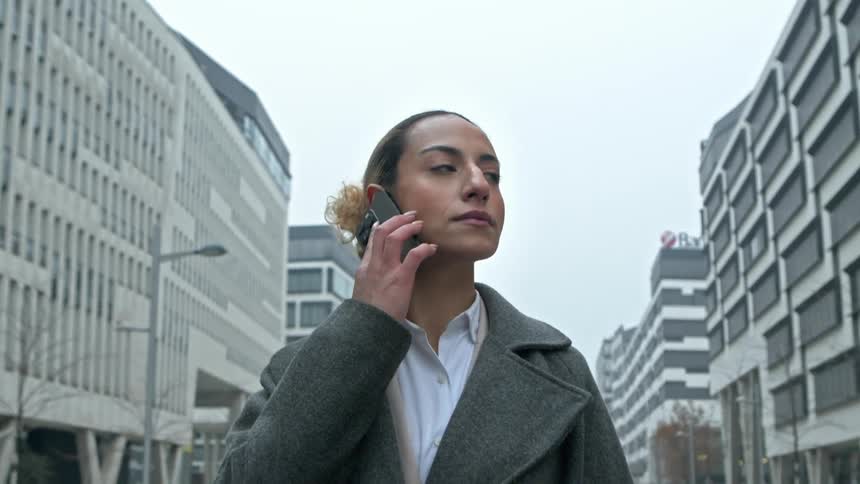 A woman in a gray coat is walking on a city street lined with modern office buildings answering a call on her smartphone